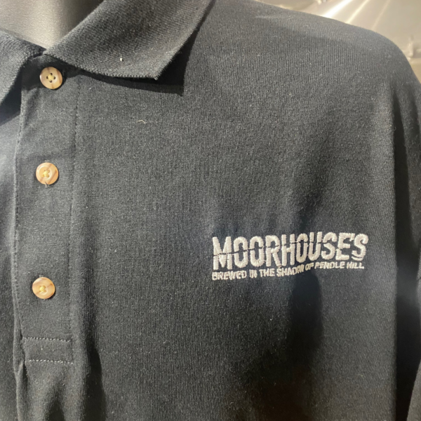 Close up of Moorhouse's Black Short Sleeve Polo Shirt with 3 brown button detail and Moorhouse's logo on left chest.